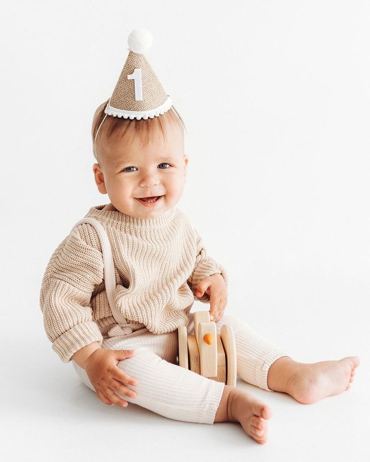 a baby sitting on the floor wearing a birthday hat and holding a wooden toy in his hands