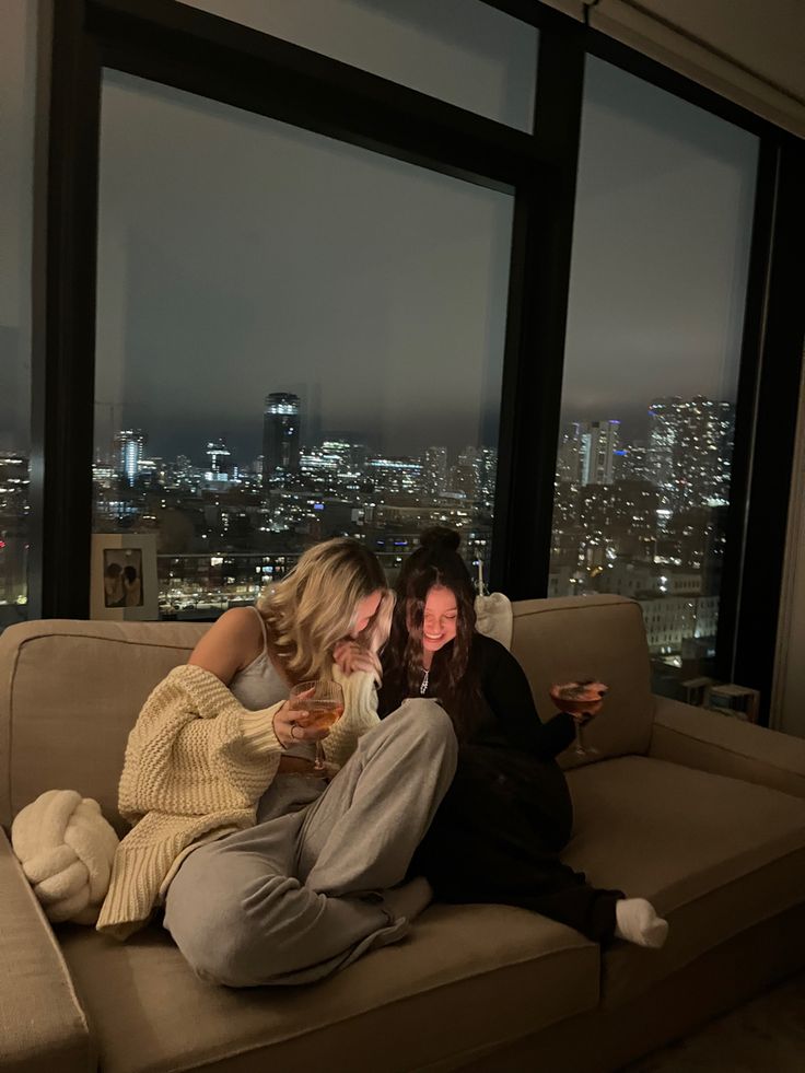 two people sitting on a couch drinking wine and looking out the window at the city