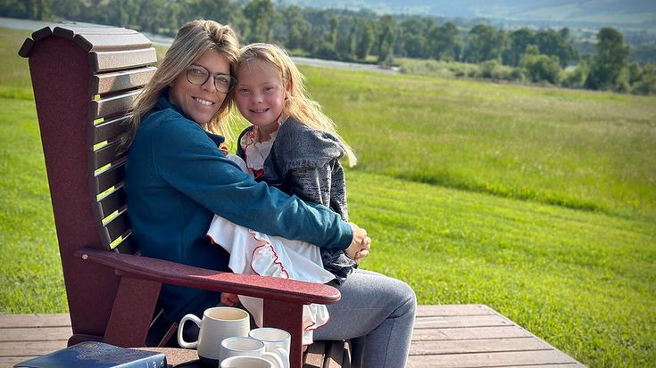 a mother and daughter sitting on a bench in the grass with their arms around each other