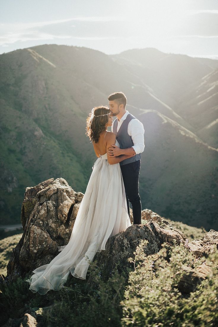 a bride and groom standing on top of a mountain in the sun with their arms around each other