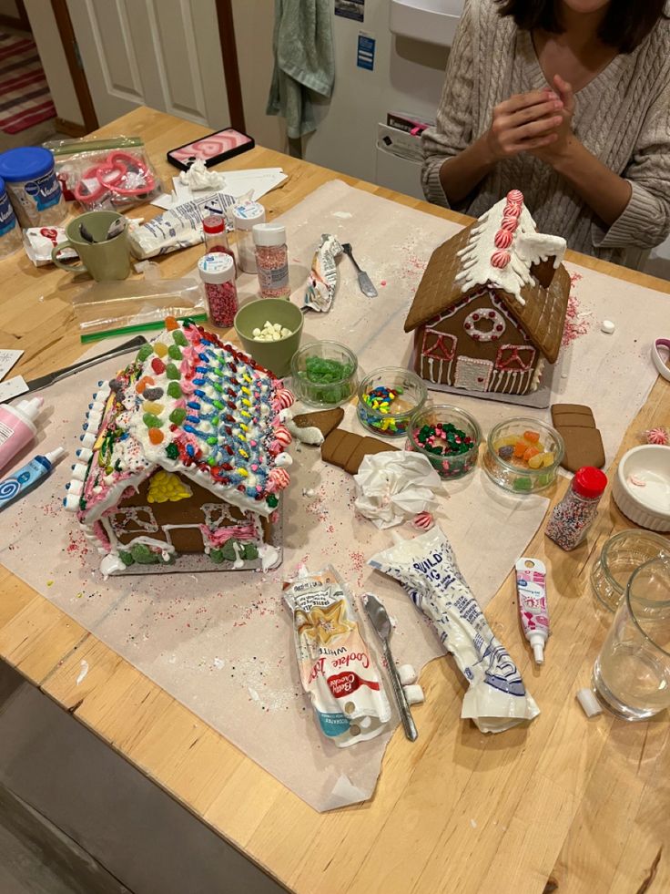 a woman sitting at a kitchen table covered in gingerbread house shaped cookies and marshmallows