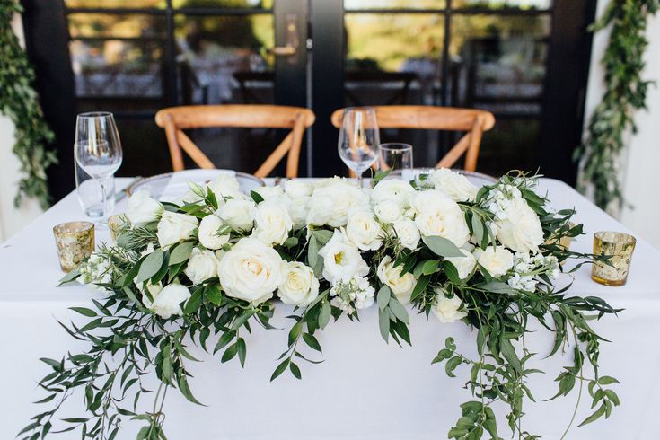 the table is set with white flowers and greenery for an elegant centerpieces