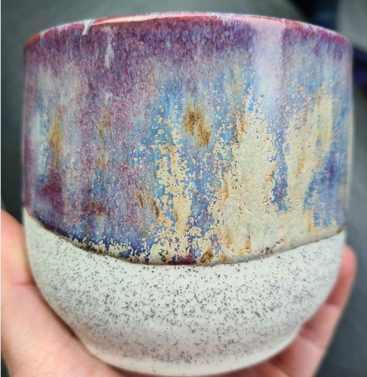 a hand holding a purple and white bowl with speckles on the outside, in front of a gray background