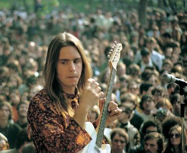 a man with long hair holding a guitar in front of a large group of people