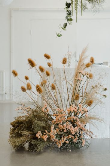 an arrangement of dried flowers and greenery in front of a white wall hanging from the ceiling