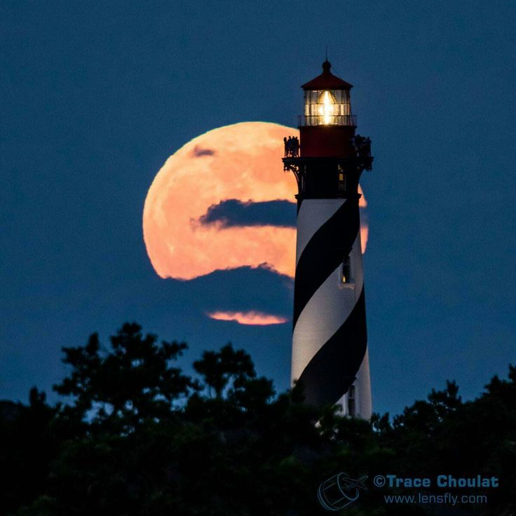 the full moon is seen behind a lighthouse in front of some trees and clouds, with a black and white stripe on it's side