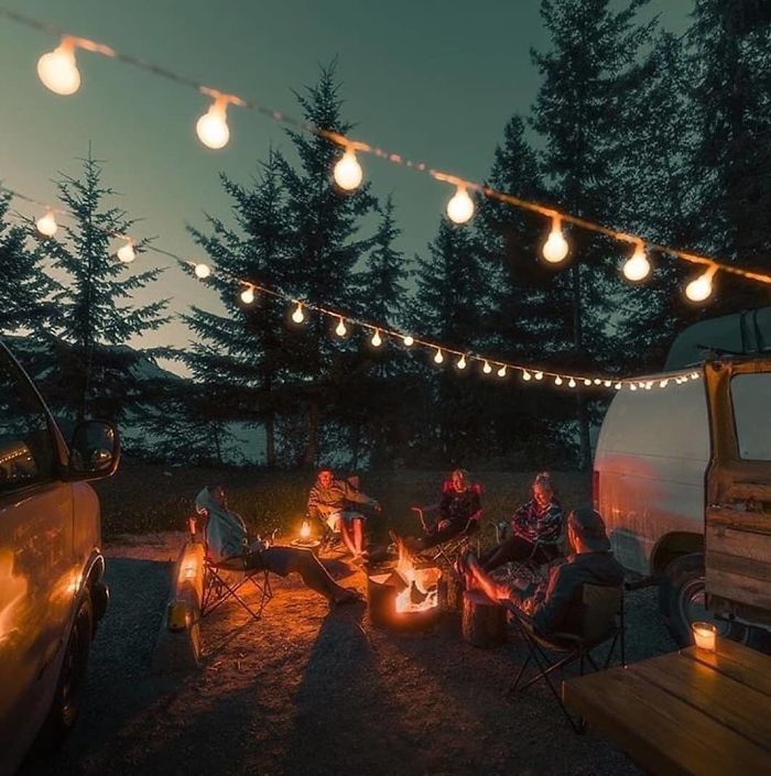 people sitting around a campfire at night with lights strung from the trees over them