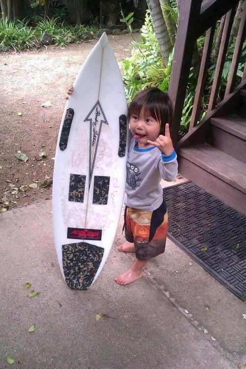 a little boy standing next to a surfboard giving the peace sign with his hand