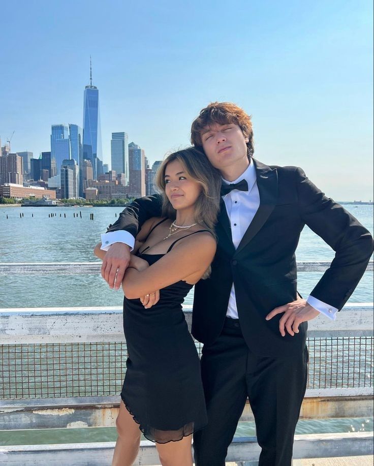 a man and woman posing for a photo in front of the water with a city skyline behind them