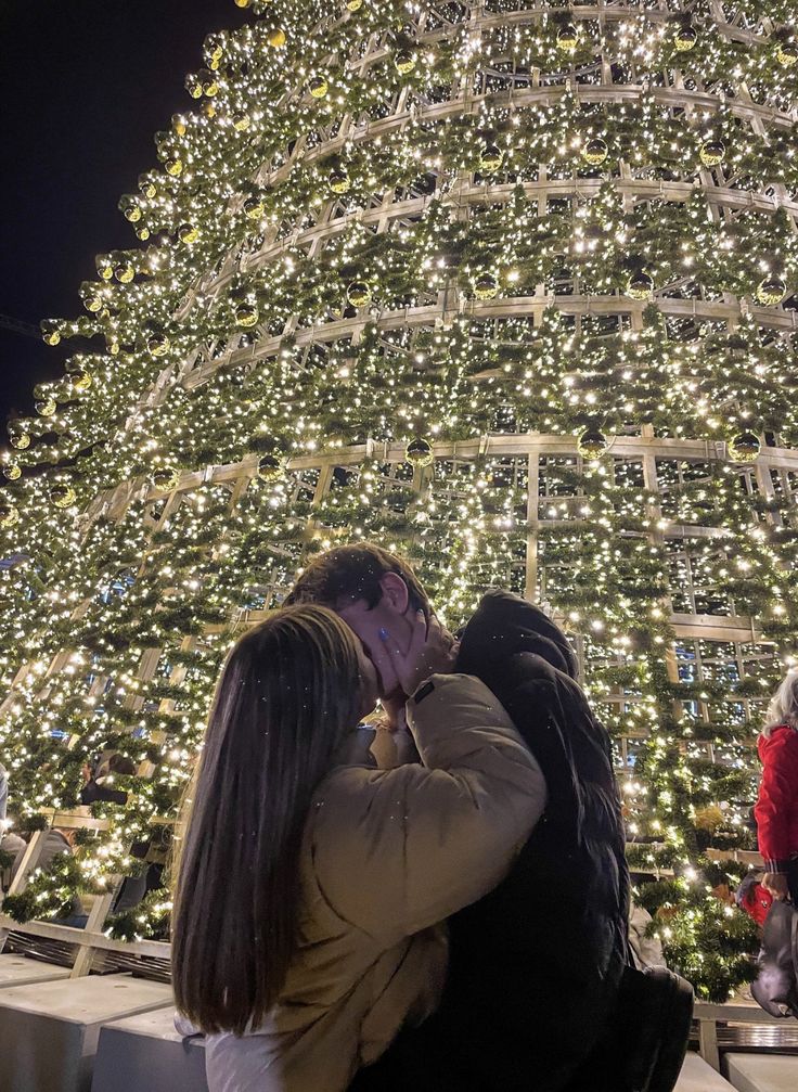 a man and woman kissing in front of a christmas tree with lights on it at night