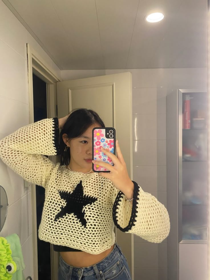 a woman taking a selfie in the bathroom with her phone and wearing a star sweater