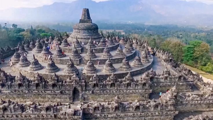 an aerial view of a temple in the middle of a forest with mountains in the background