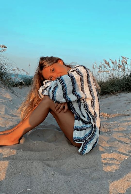 a woman sitting in the sand with her legs crossed and wearing a striped towel over her shoulders