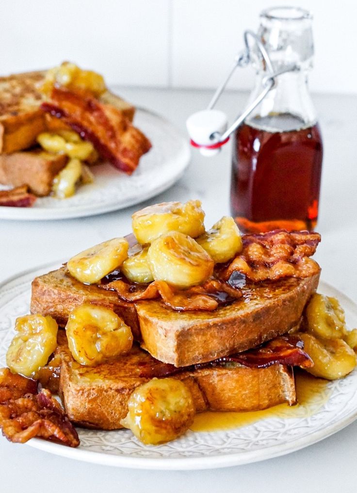 two white plates topped with french toast covered in bananas and syrup next to a bottle of syrup