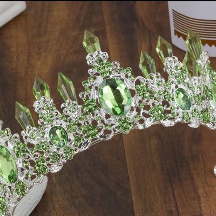 a tiara with green stones on it sitting on a wooden table next to a cup