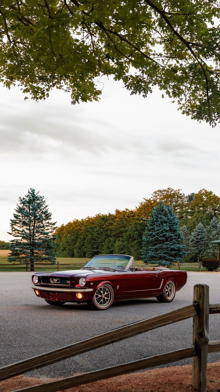 a classic car parked in a parking lot next to a wooden fence and some trees