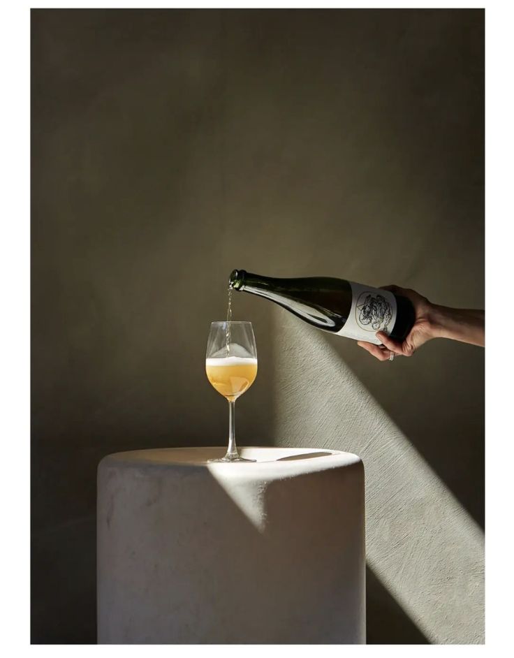 a person pouring wine into a glass on top of a white tablecloth with the shadow of a wall behind it