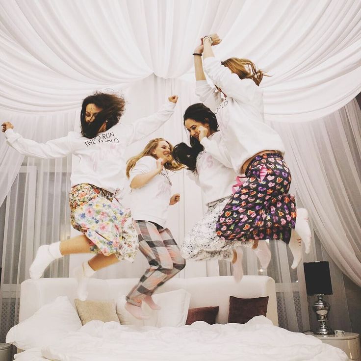 four girls jumping in the air on a bed with white drapes and curtains behind them