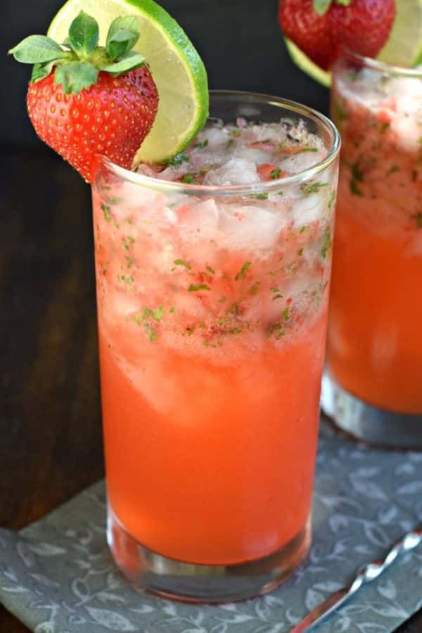 two glasses filled with drinks and garnished with strawberries, limes and mint