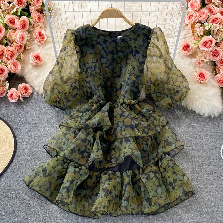 Immerse yourself in the whimsy of our Enchanted Forest Tiered Ruffle Dress, where elegance meets a touch of the untamed. This dress features a captivating camouflage print in shades of forest green and earthy browns, reminiscent of a woodland escape. The sheer, puffed bishop sleeves add a dash of romance and drama, while the lightweight fabric ensures a flowy, ethereal movement with every step you take.The dress boasts a cinched waistline that gracefully gives way to a tiered ruffle skirt, creat Chic Dark Green Midi Dress For Summer, Spring Party Dark Green Mini Dress, Summer Party Dark Green Midi Dress, Green Mini Dress With Ruffle Hem, Fall Chiffon Dress For Garden Party, Elegant Dark Green Mini Dress, Dark Green Mini Dress For Summer, Chic Dark Green Midi Dress For Spring, Elegant Chiffon Mini Dress With Floral Print