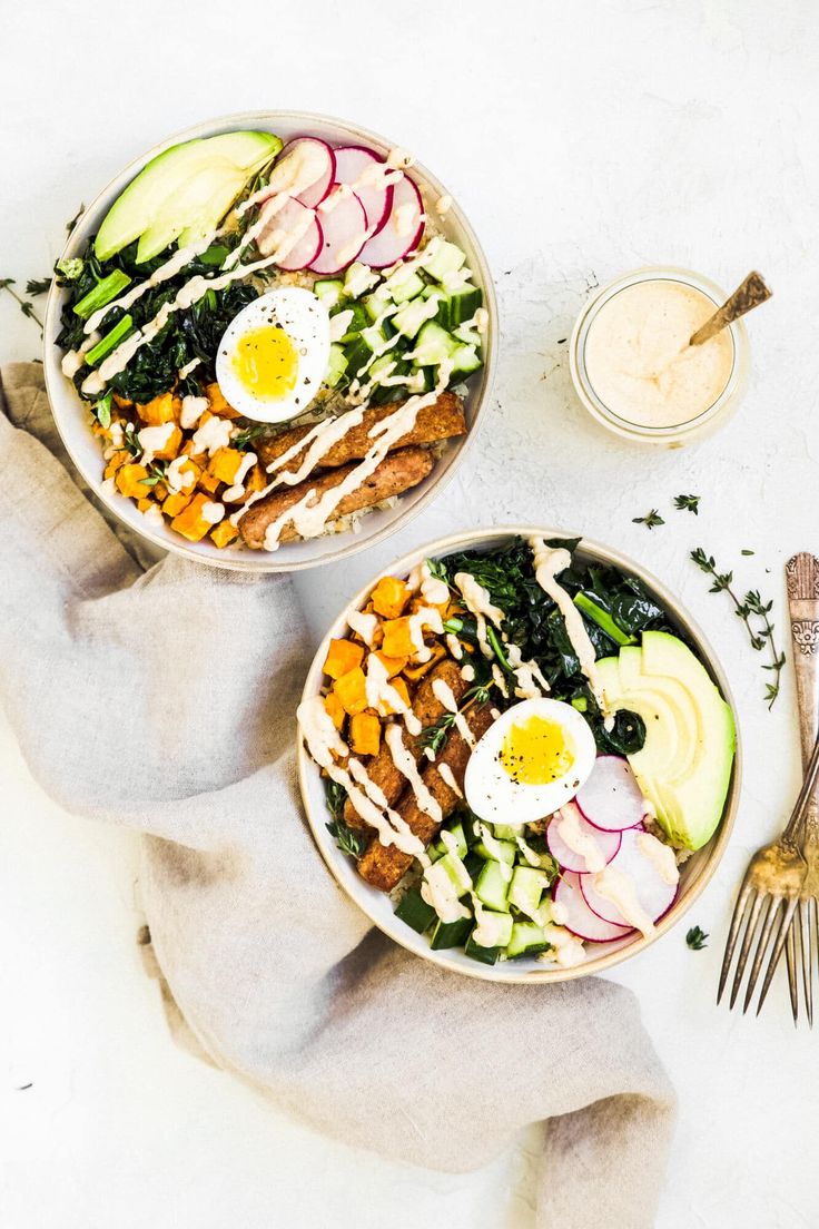 two bowls filled with vegetables and eggs on top of a white surface next to a fork