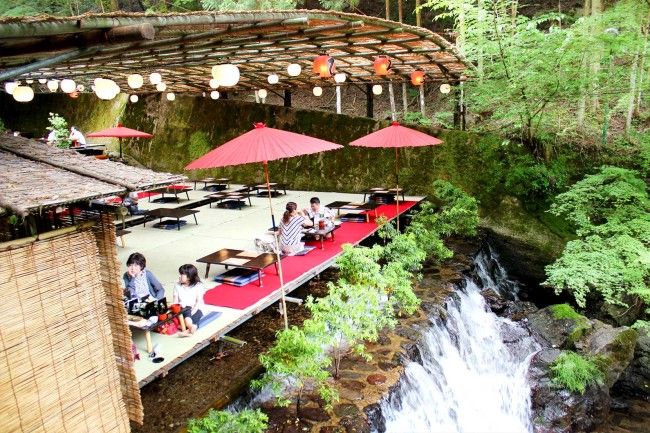 people sitting at tables near a waterfall under red umbrellas in the forest with lights hanging above them