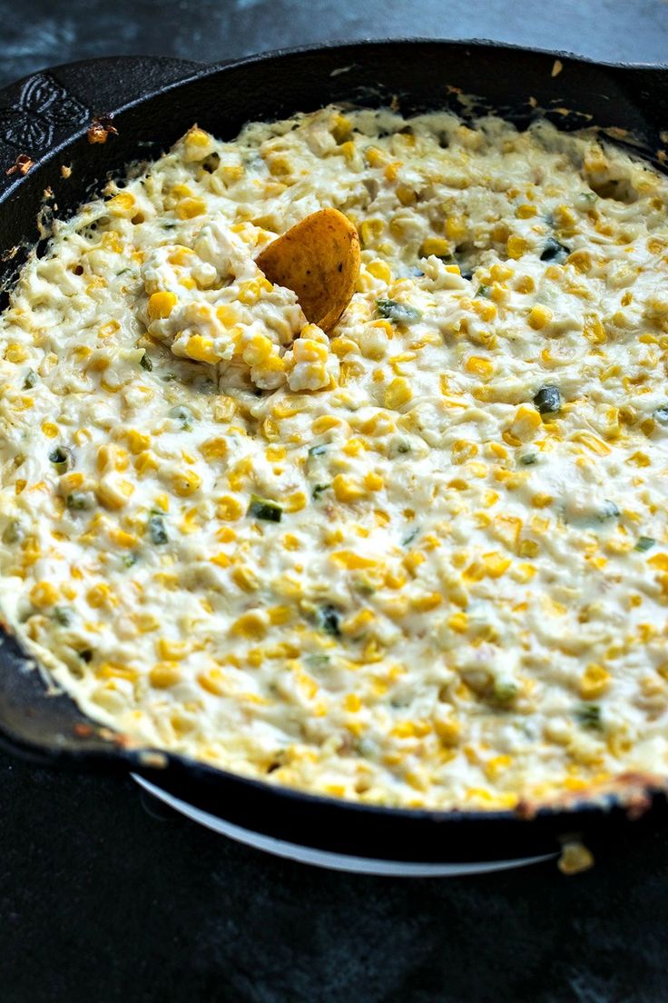 a skillet filled with corn and cheese on top of a table next to a wooden spoon