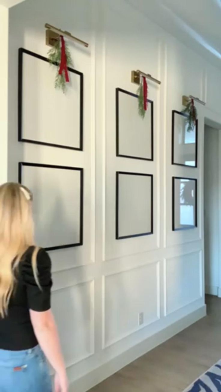 a woman standing in front of a white wall with black frames on it and christmas decorations