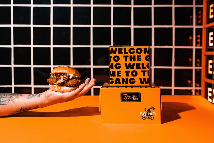 a hand holding a burger in front of a box with the words welcome to the gang on it