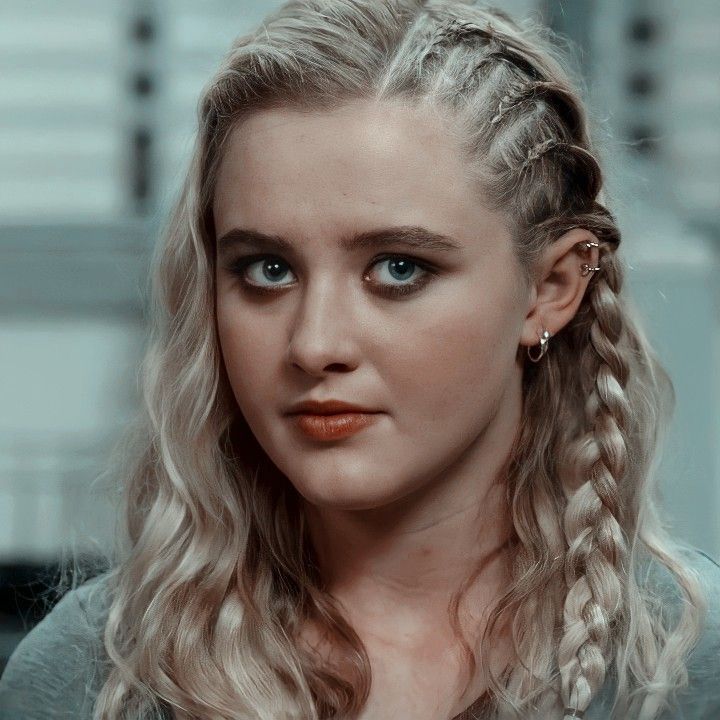 a woman with long blonde hair and blue eyes looks into the camera while wearing braids