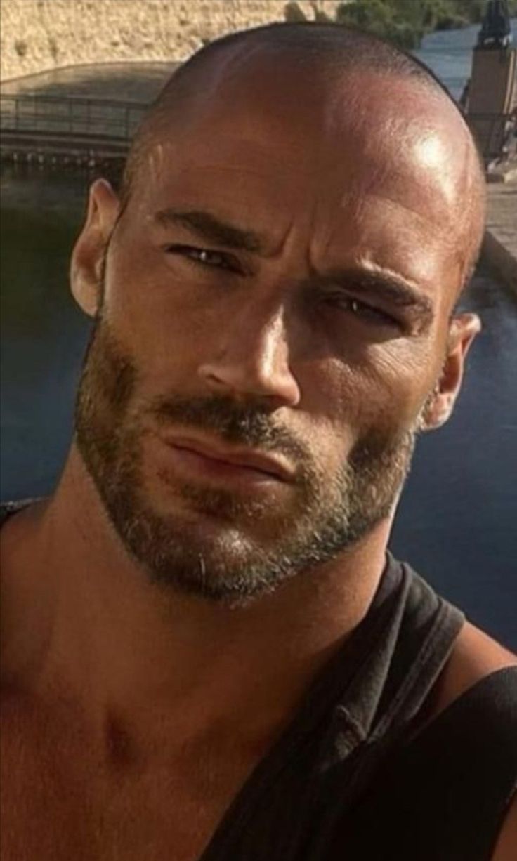 a man with a bald head and no shirt is looking at the camera while standing next to a body of water