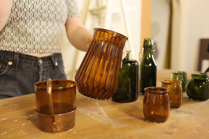 a woman standing next to some brown glass cups and vases on a wooden table