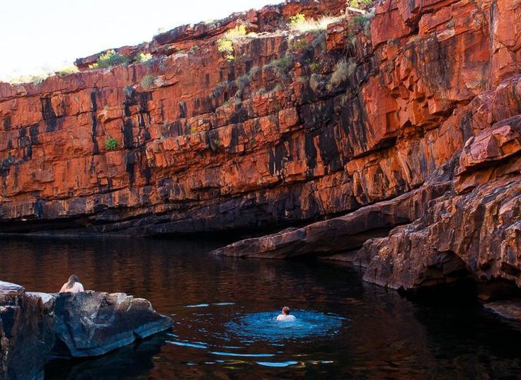 there is a man swimming in the water near some red rocks and cliffs with trees on top