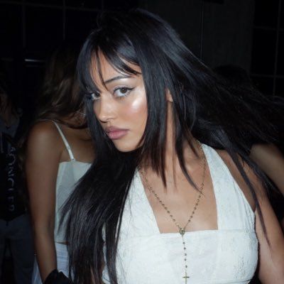 a woman with long black hair standing in front of other women wearing white dresses and necklaces