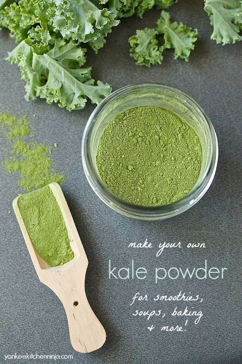 kale powder in a glass bowl next to a wooden spoon and leafy greens