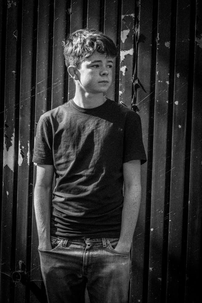 a young man standing in front of a wooden fence with his hands in his pockets