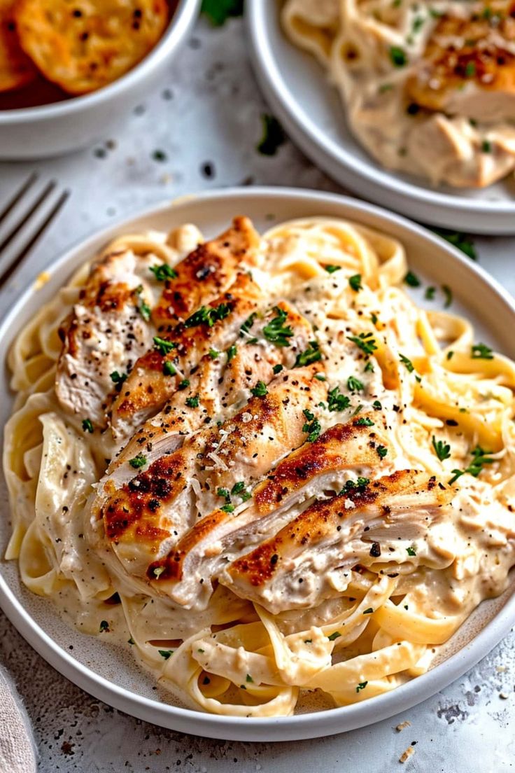 two white plates filled with pasta and chicken alfredo on top of each other next to silverware