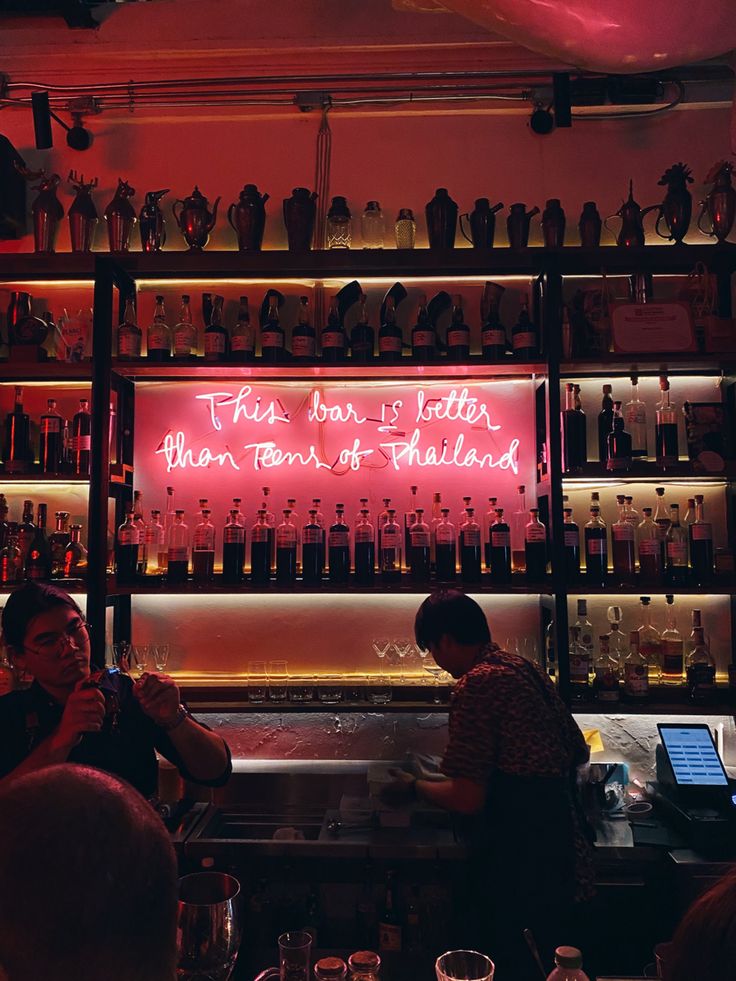 people are sitting at a bar with neon signs on the wall and bottles behind them