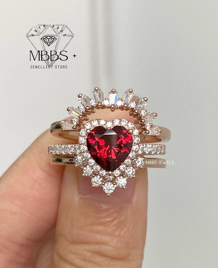 a woman's hand holding a heart shaped ring with diamonds around it and a red stone in the middle