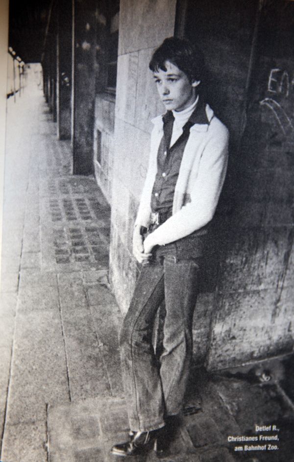 a young man leaning against a wall with his hands in his pockets, wearing a suit and tie