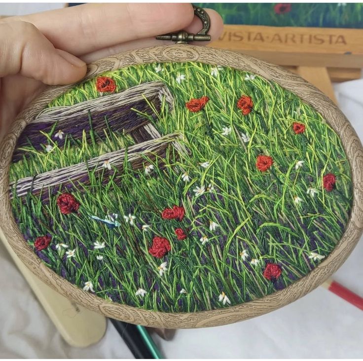 someone is holding up a small embroidery art piece with grass and red flowers on it