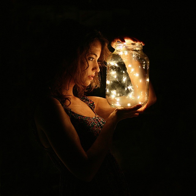 a woman holding a jar filled with lights in the dark, while looking at it