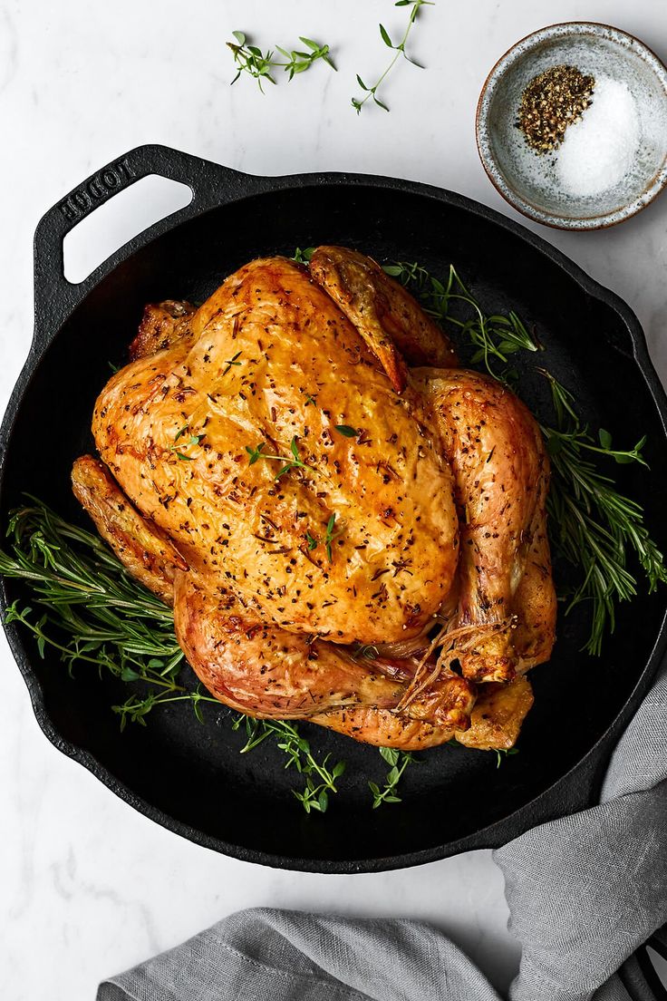 a roasted chicken in a cast iron skillet with herbs and seasoning on the side