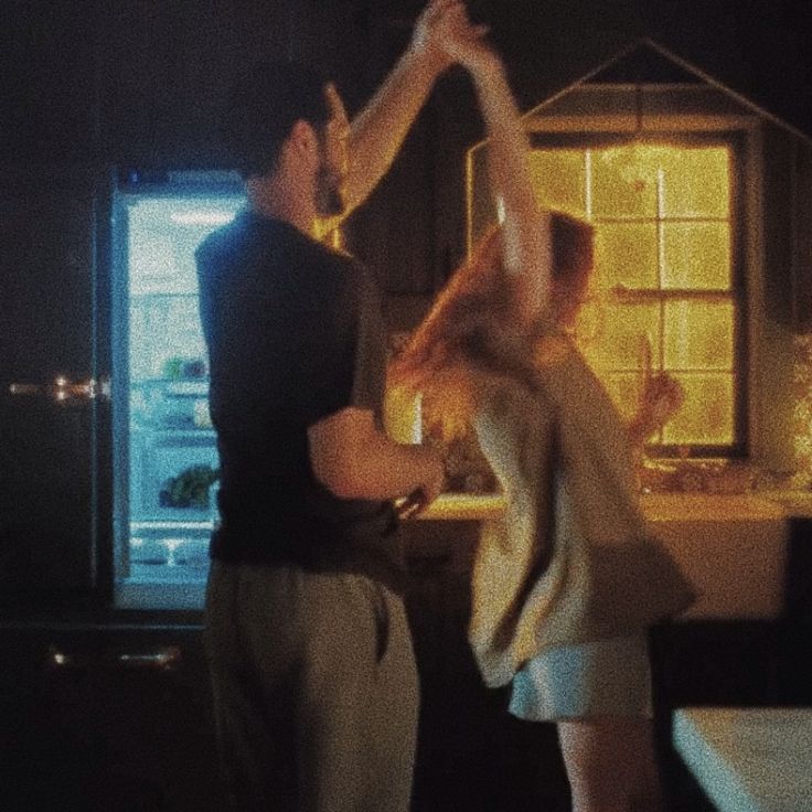 two people are dancing in front of a house at night with their hands up to the ceiling