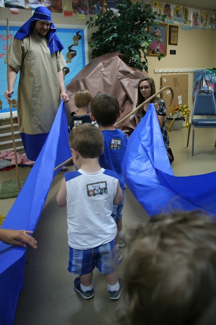 a group of young children holding blue kites in a room with adults and children