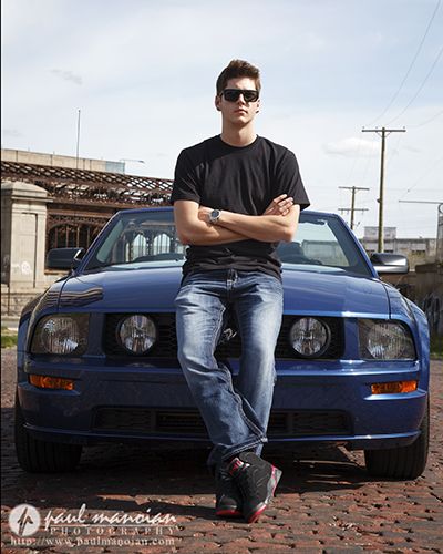 a young man sitting on the hood of a blue sports car with his arms crossed