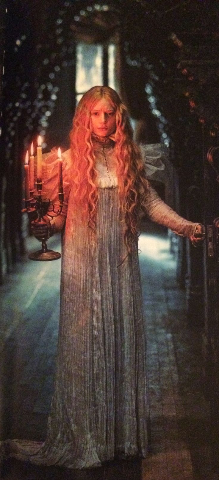 a woman with long blonde hair standing in an alley next to candles and looking at the camera