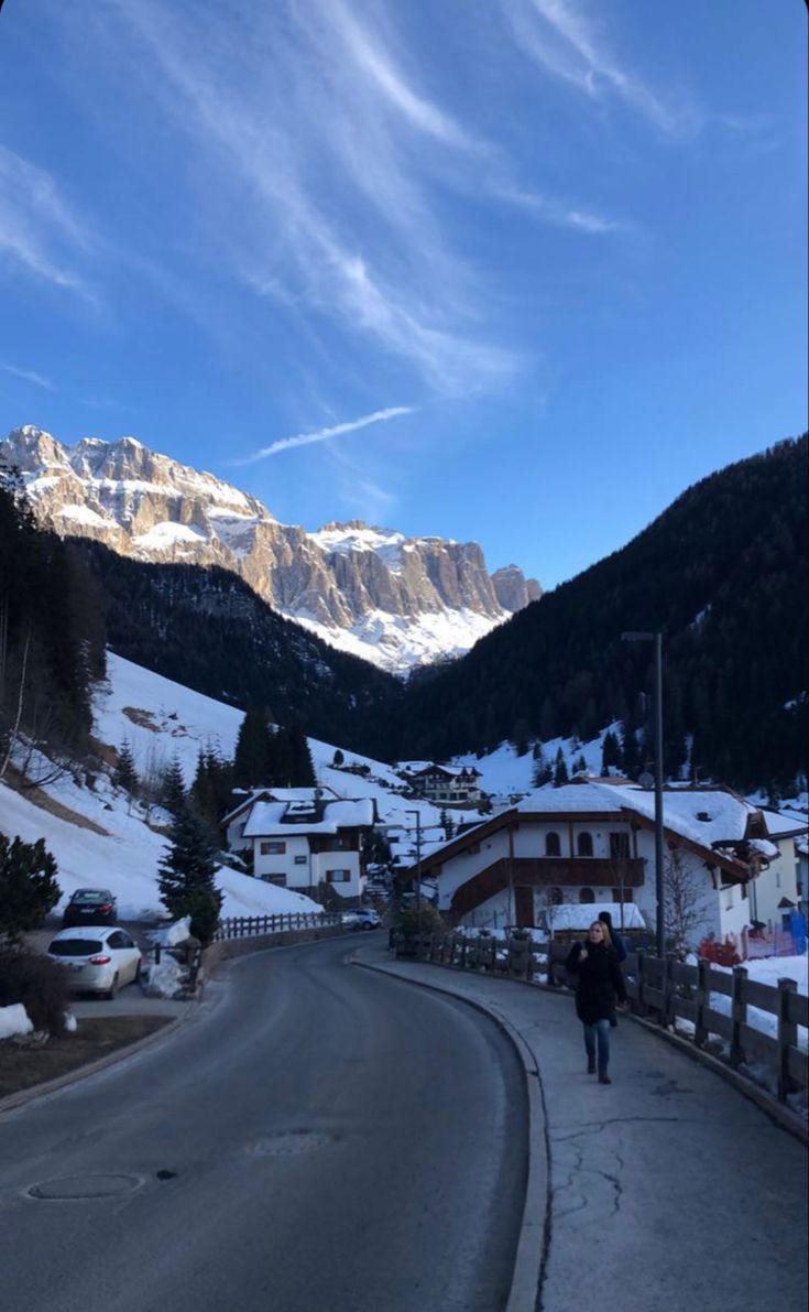 a person walking down the road in front of some snow covered mountains and houses on either side