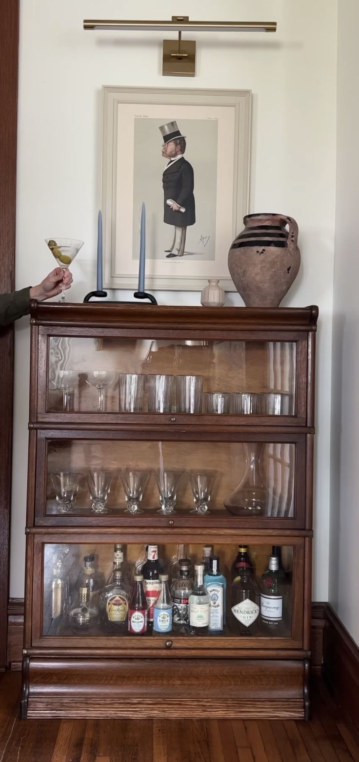 a wooden cabinet filled with glasses and bottles on top of a hard wood floor next to a painting