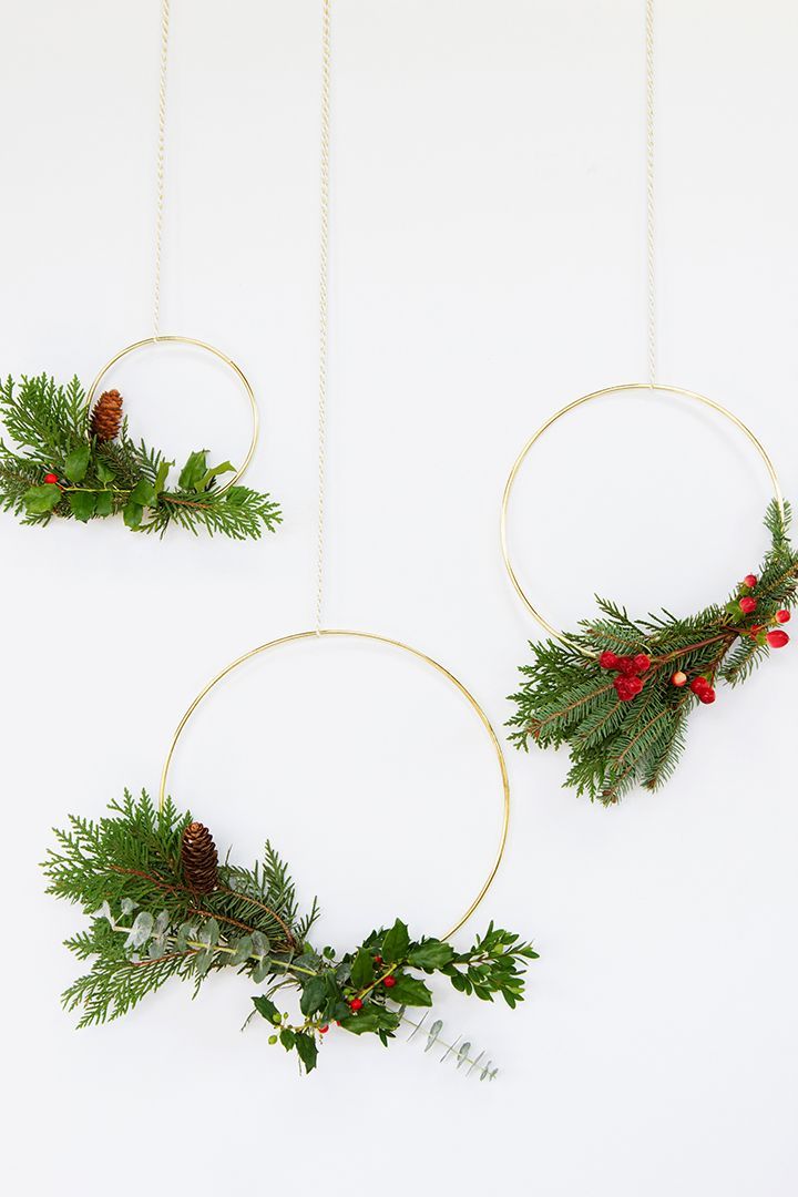 two christmas wreaths hanging from gold hoop with holly and pine cones on white background